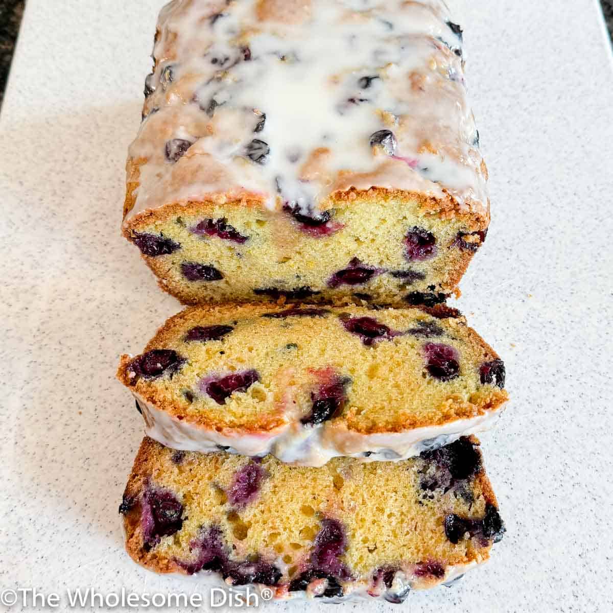 Sliced glazed lemon blueberry bread on a white cutting board.