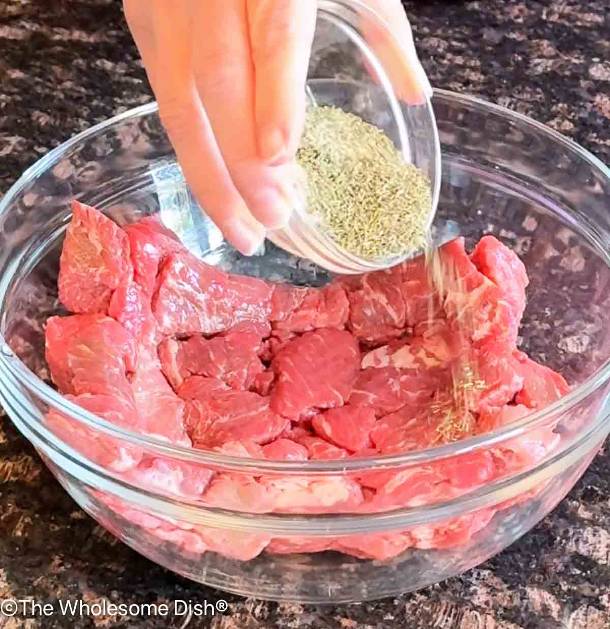 Pouring seasonings over raw beef stew meat in a glass bowl.